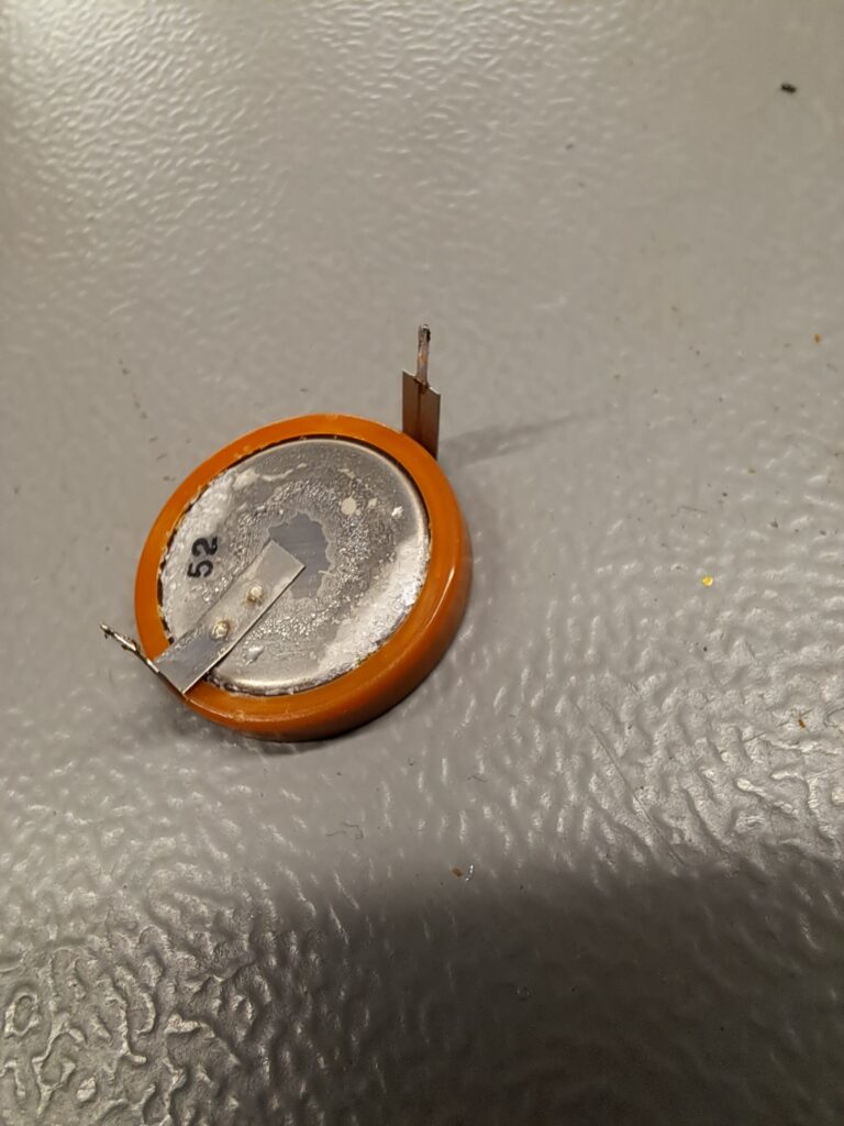 Verwijderde soldeerbare knoopcel (CMOS-batterij) met kristallijne lekkage op het contactoppervlak — corrosief materiaal dat omliggende printbanen snel aantast