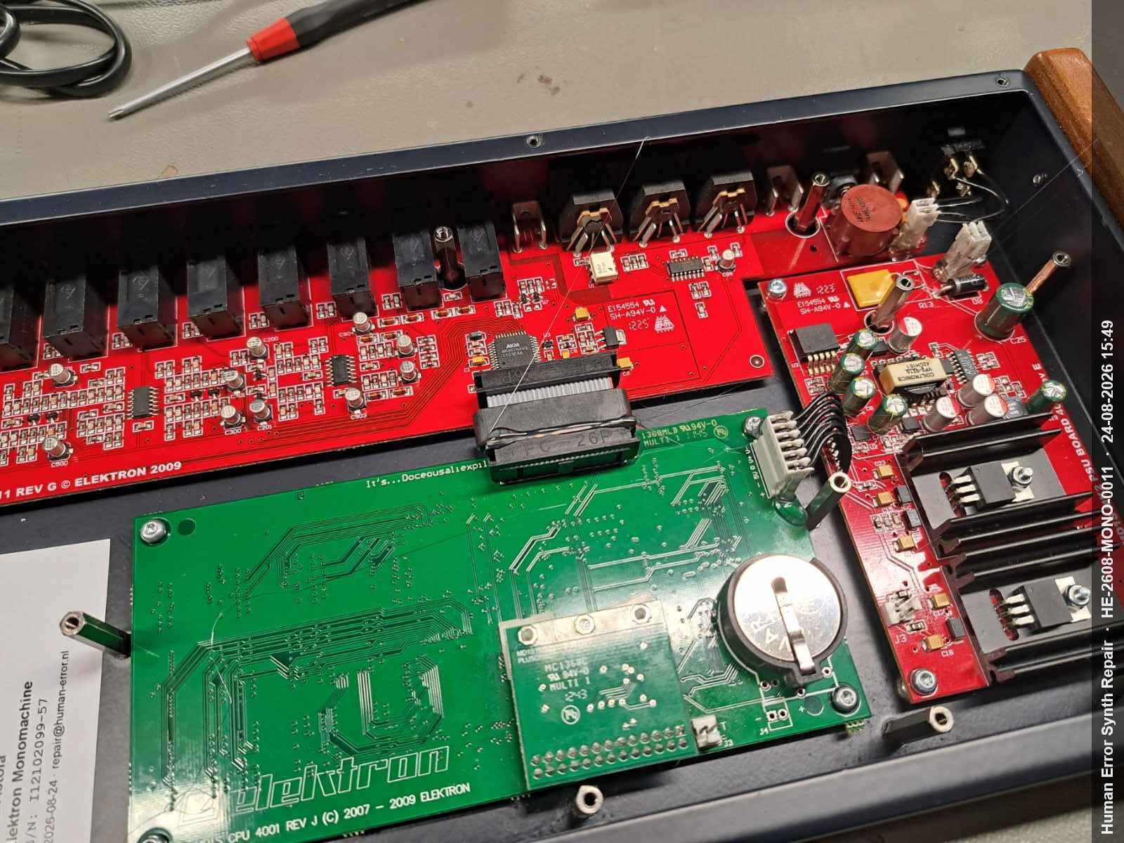 Geopende Monomachine met groen CPU-board, rood IO-board, PSU-board en nieuwe CR2450-batterij zichtbaar naast elkaar.